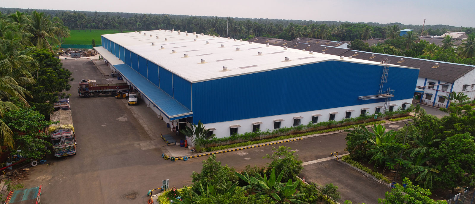 Large industrial warehouse with blue and white exterior, surrounded by greenery
